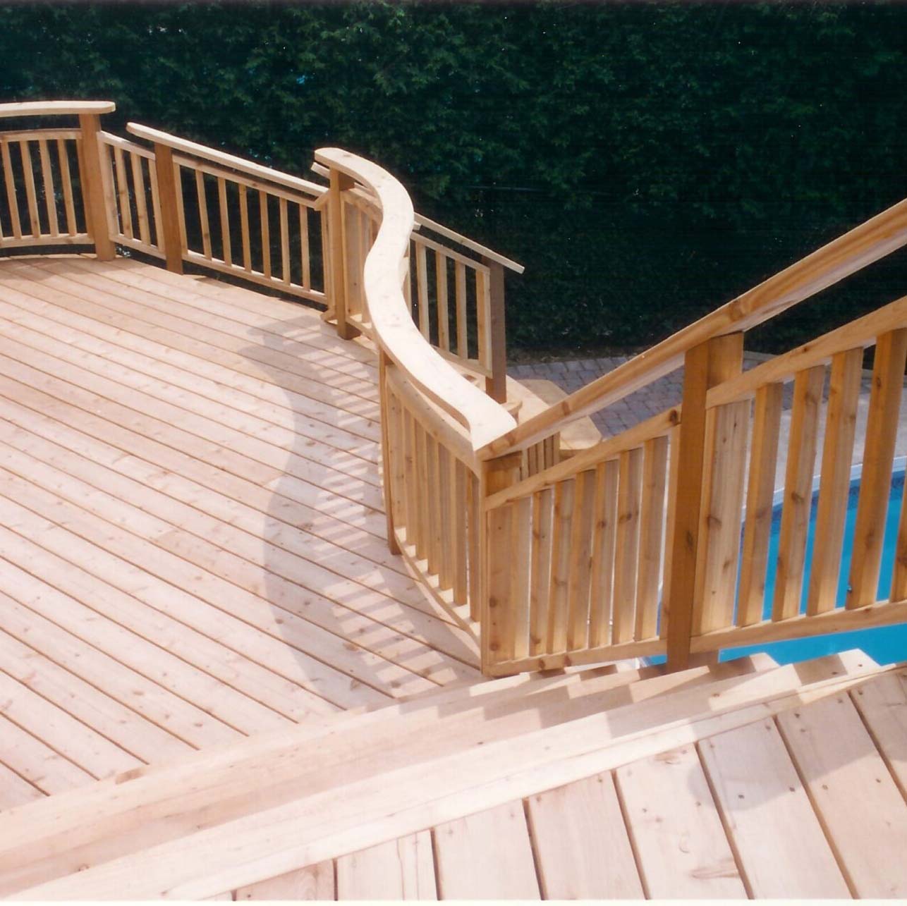 A wooden deck with curved railing overlooks a pool and green hedge, showcasing craftsmanship and inviting outdoor relaxation space. No people are present.