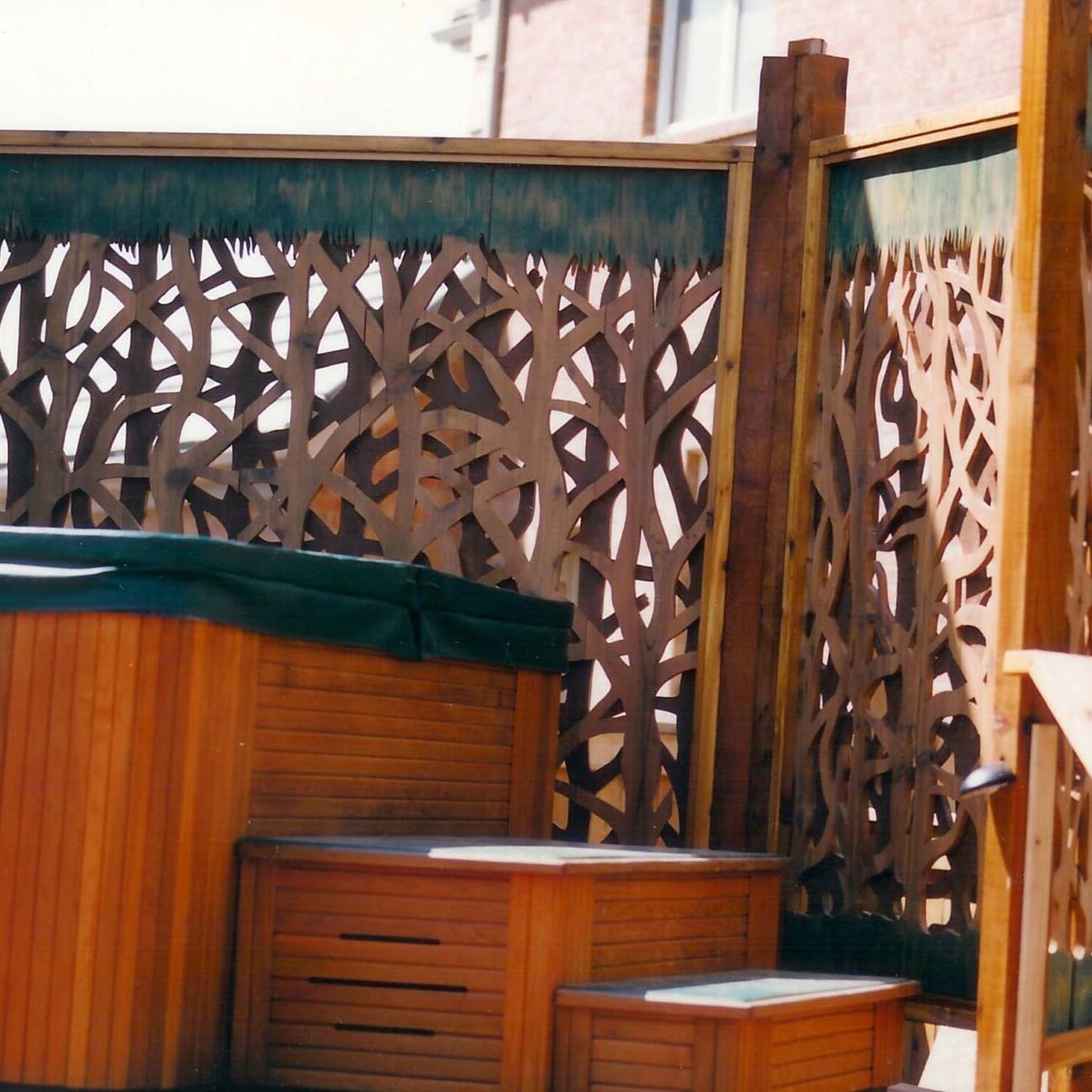 A wooden hot tub with a decorative privacy screen, featuring intricate latticework, is set against a brick wall in a backyard.