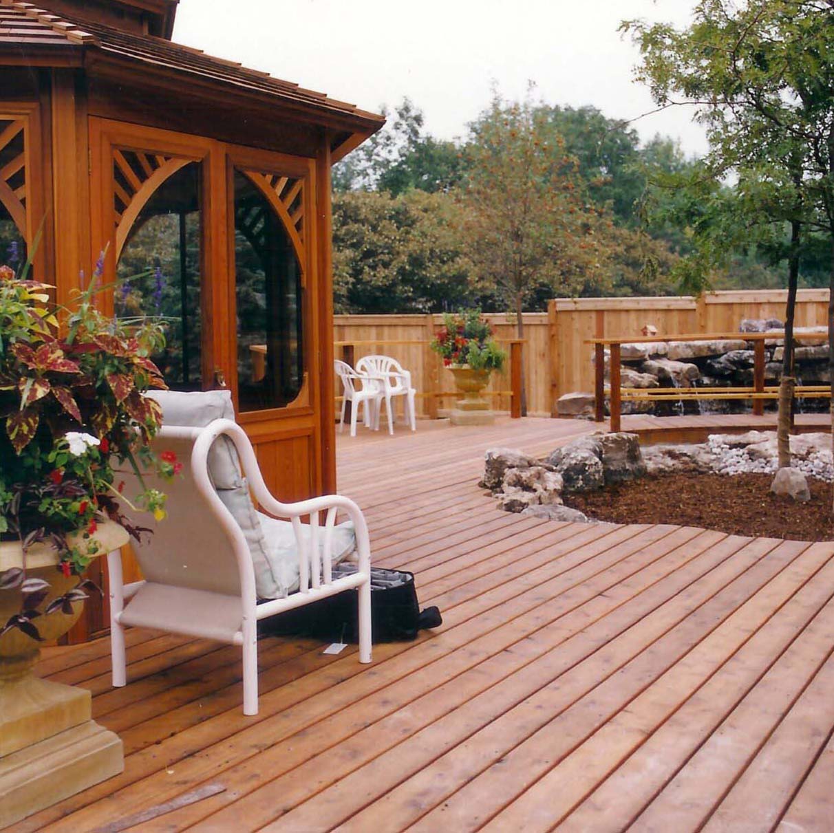 A wooden deck features a gazebo, seating area, potted plants, and a small rock garden, surrounded by a wooden fence and greenery.