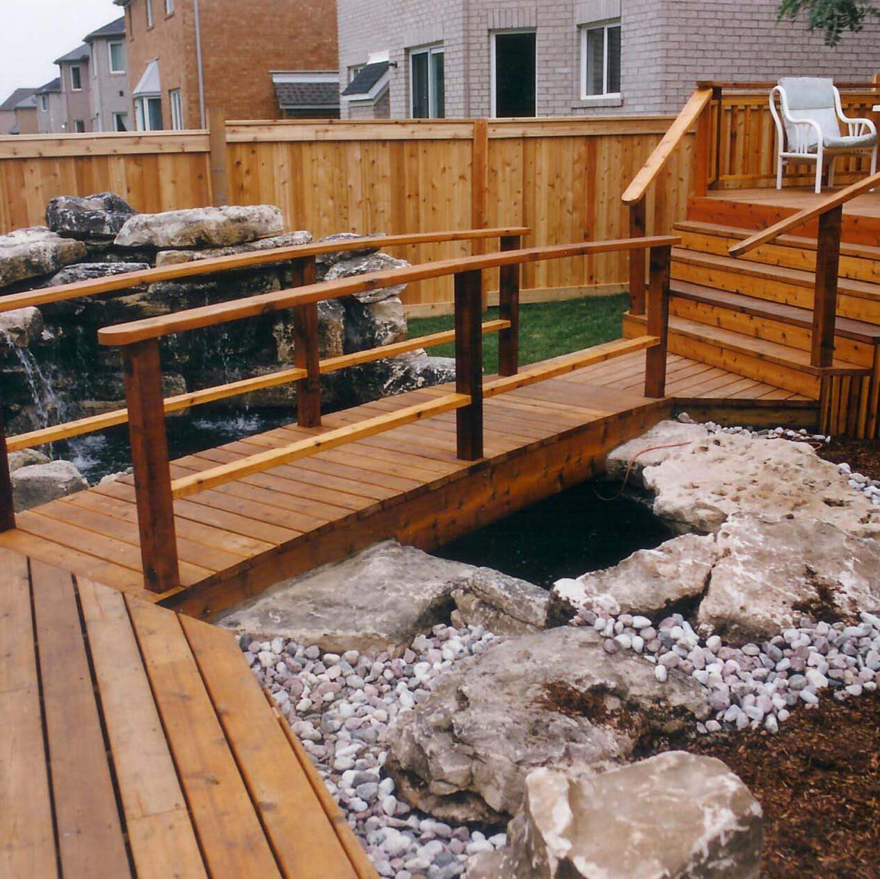 A wooden deck with a bridge spans over a small rock garden and pond, surrounded by a fenced yard and neighboring houses.