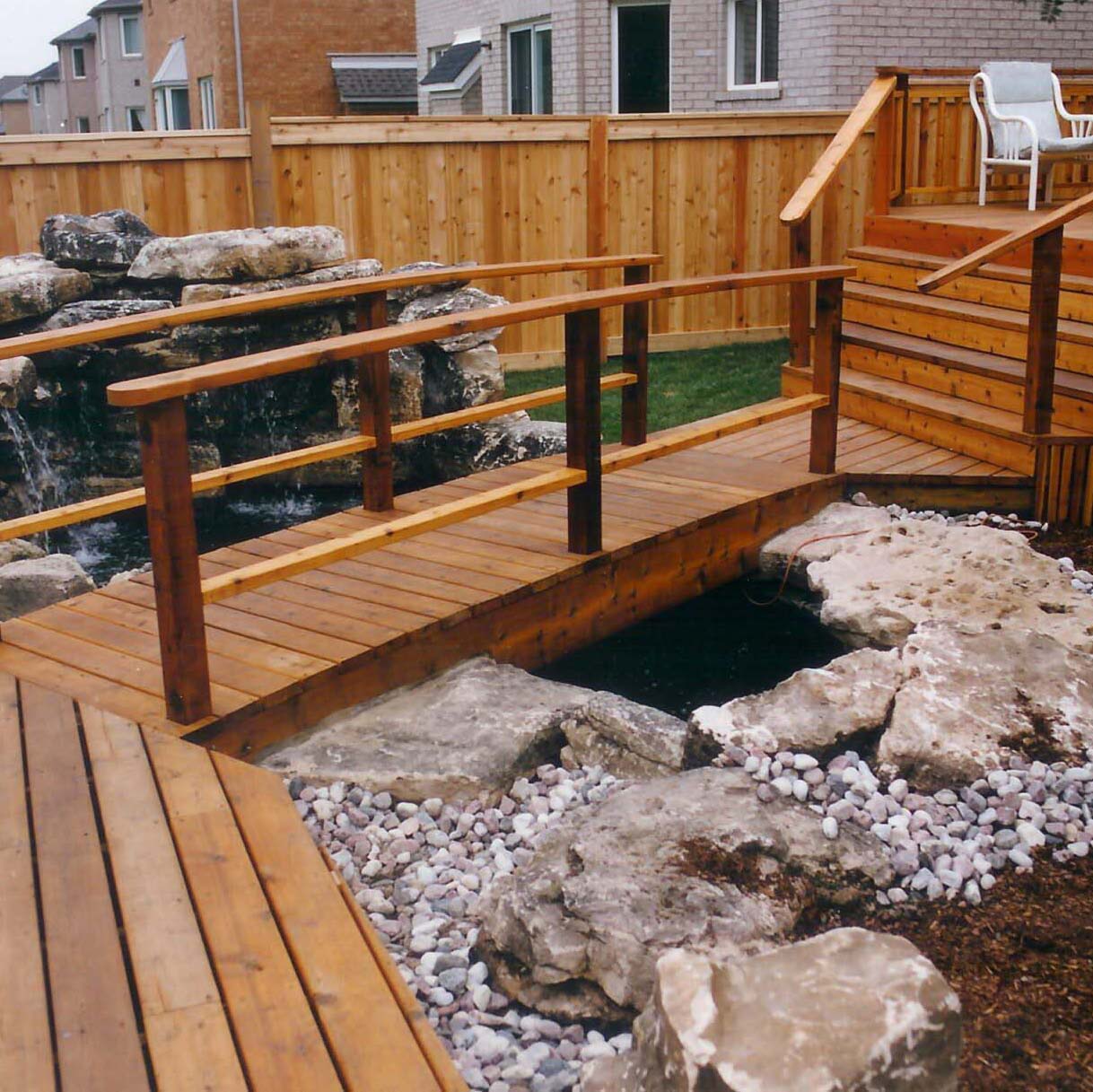 A wooden deck with railings overlooks a neatly arranged rock garden and koi pond, surrounded by a privacy fence.