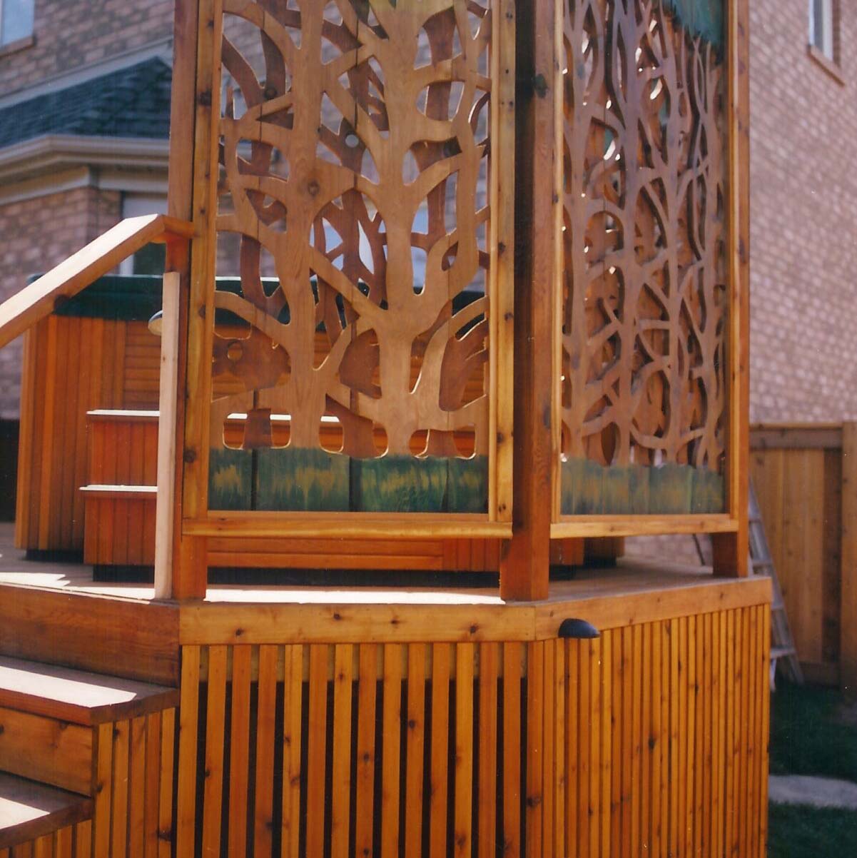 Wooden deck structure with ornate cutout panels, situated in a residential backyard setting, next to a brick house. No landmarks visible.