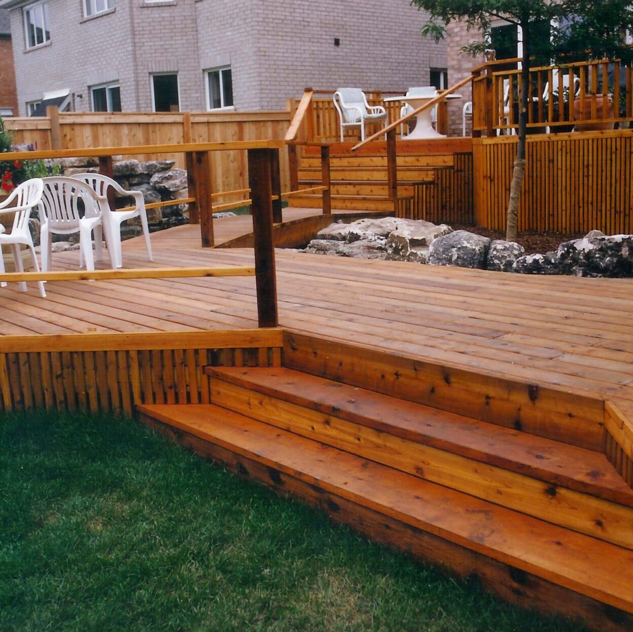 A spacious wooden deck with multiple levels, white chairs, and a fence, adjacent to a modern brick building and grassy area.