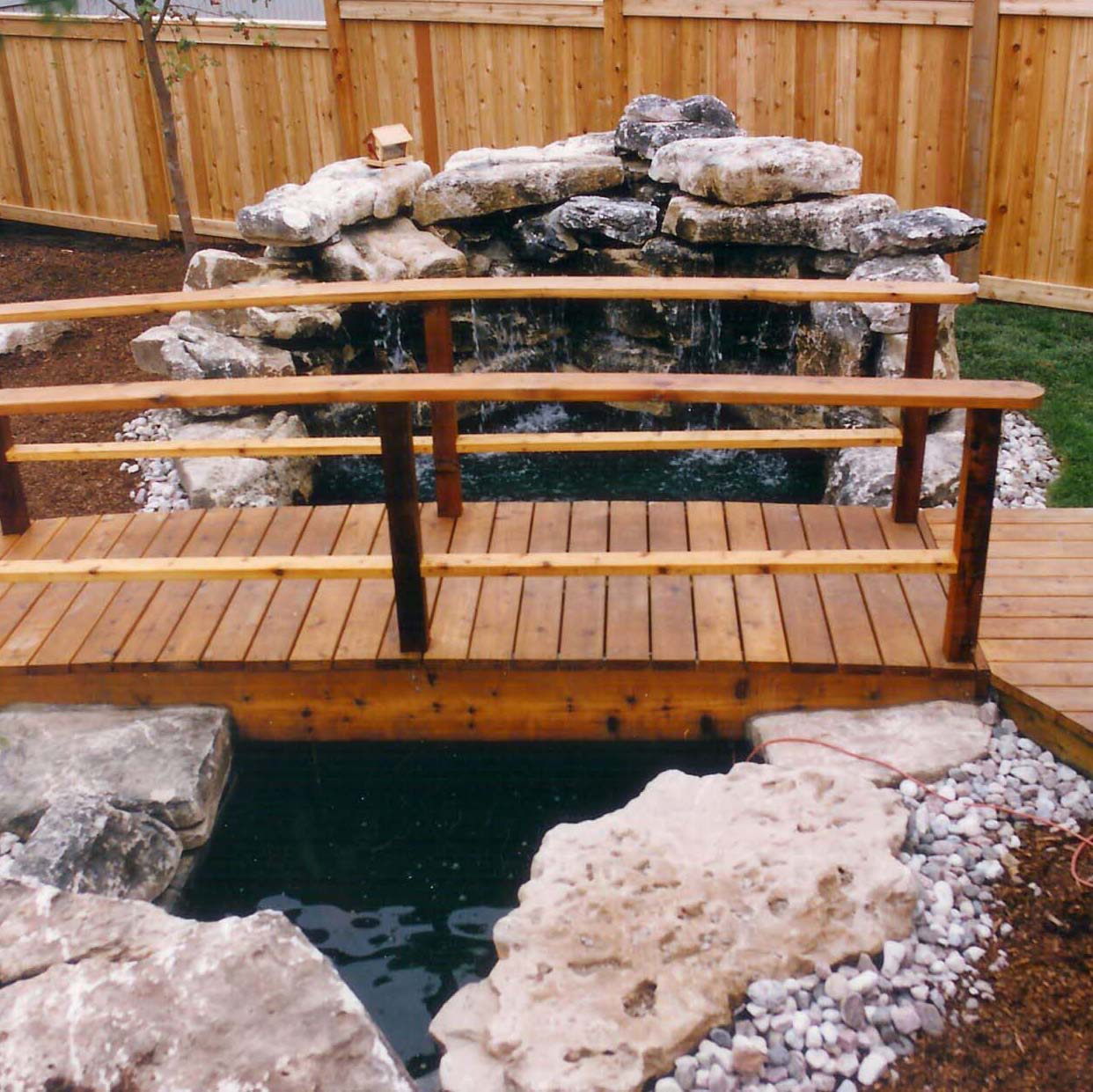 A wooden footbridge stretches over a small artificial pond with stone edges, surrounded by a wooden fence, in a landscaped backyard.