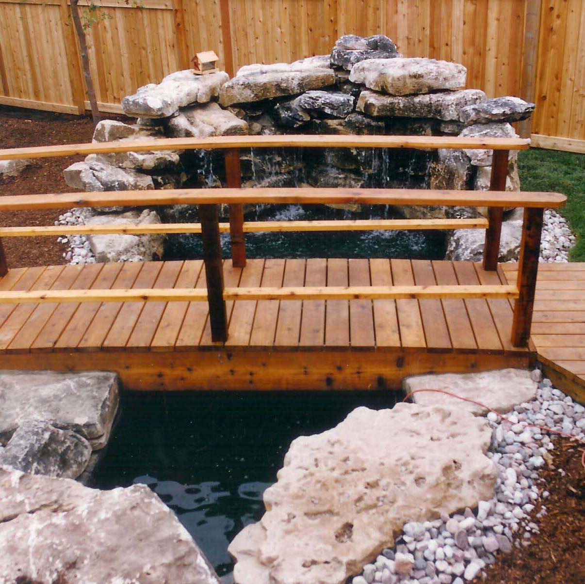 A wooden bridge spans a small pond, surrounded by rocks and a wooden fence, with a waterfall feature in a landscaped area.