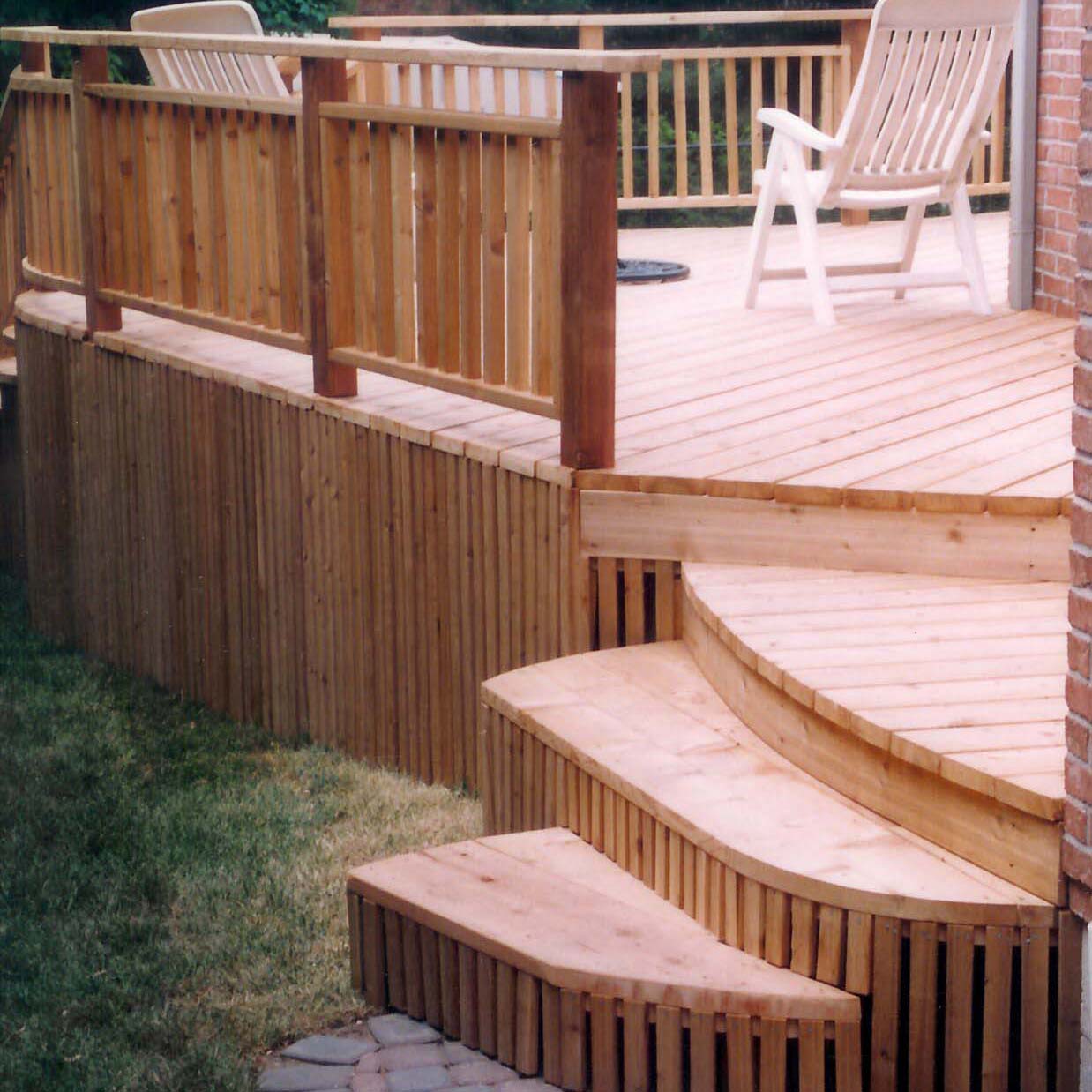 Wooden deck with railings and curved steps, featuring patio chairs. The deck is attached to a brick wall, surrounded by grass.