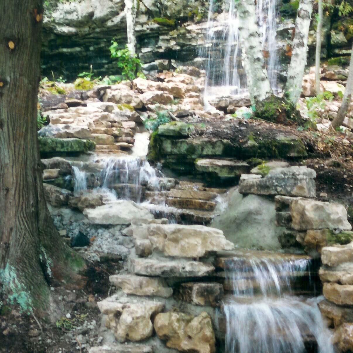 A serene waterfall cascades over layered rocks in a lush forest setting, surrounded by trees and greenery, creating a peaceful natural scene.