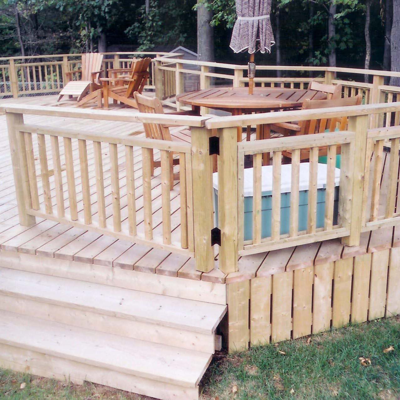 A wooden deck with railings features several wooden chairs, a round table with an umbrella, surrounded by lush green trees in the background.