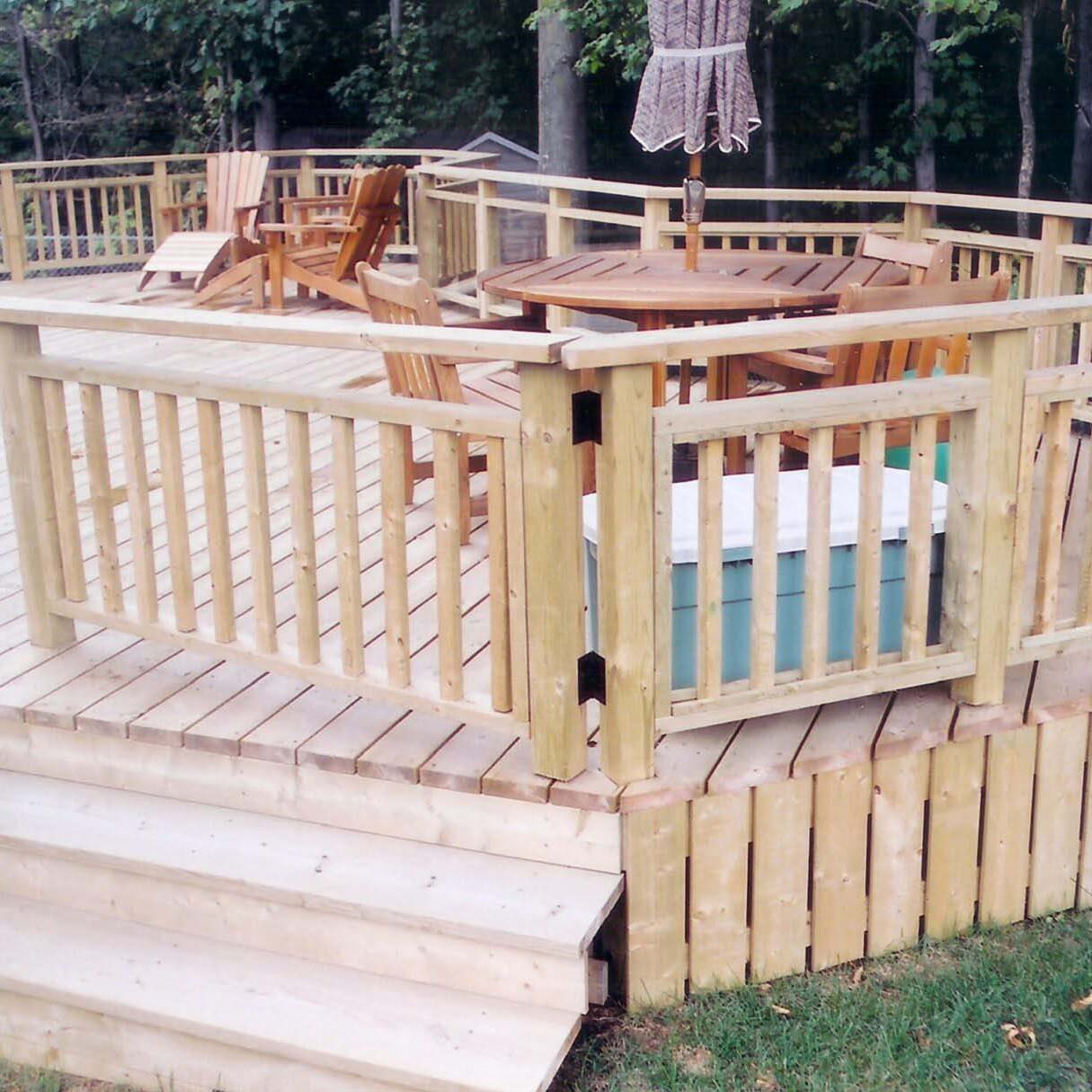 A wooden deck features Adirondack chairs, a round table, and railing, set amidst trees, providing a cozy outdoor space for relaxation.