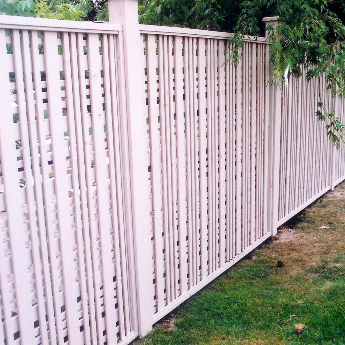 Tall white picket fence in a garden. Green trees overhang the fence. Grass covers the ground, creating a peaceful, private outdoor space.