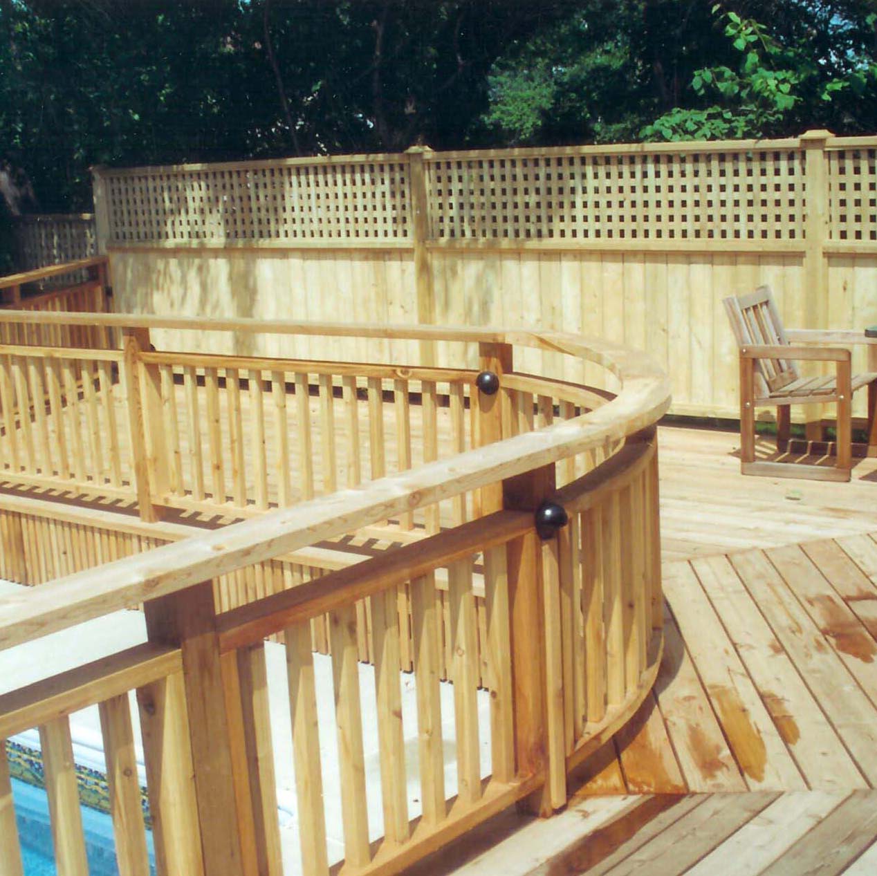 A wooden deck features curved railings and an empty chair, surrounded by a lattice fence. Trees provide a lush green backdrop.