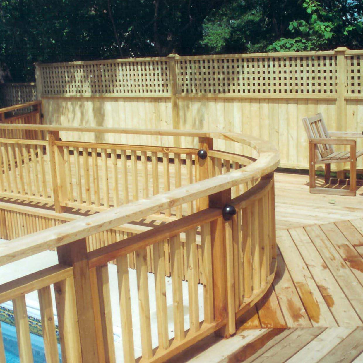 A wooden deck surrounds a swimming pool, featuring a curved railing. A solitary chair rests in the corner, with a wooden fence backdrop.