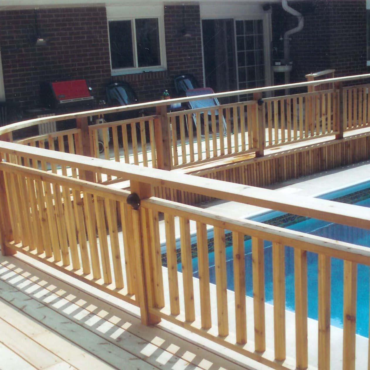 Wooden deck with a railing surrounds an outdoor swimming pool. Brick building with glass doors and patio furniture in the background. No landmarks visible.