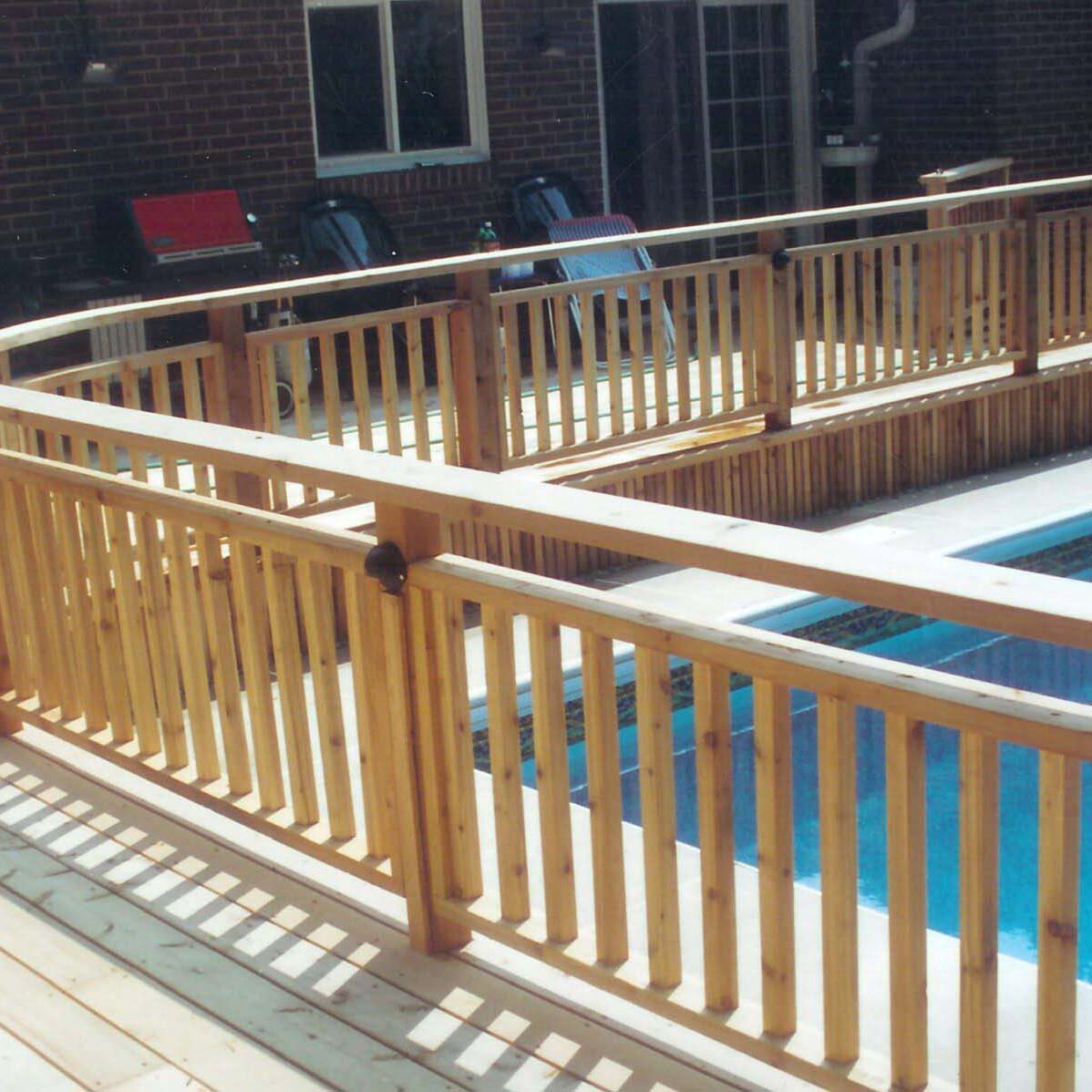 A wooden deck with a safety railing surrounds a pool. Chairs and a barbecue grill are positioned near a brick building.