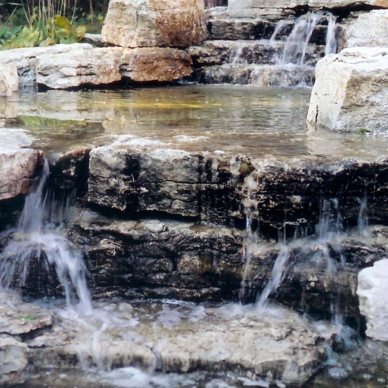 A small, tiered rock waterfall gently flows over mossy stones. Surrounding foliage adds natural beauty to the tranquil outdoor scene.