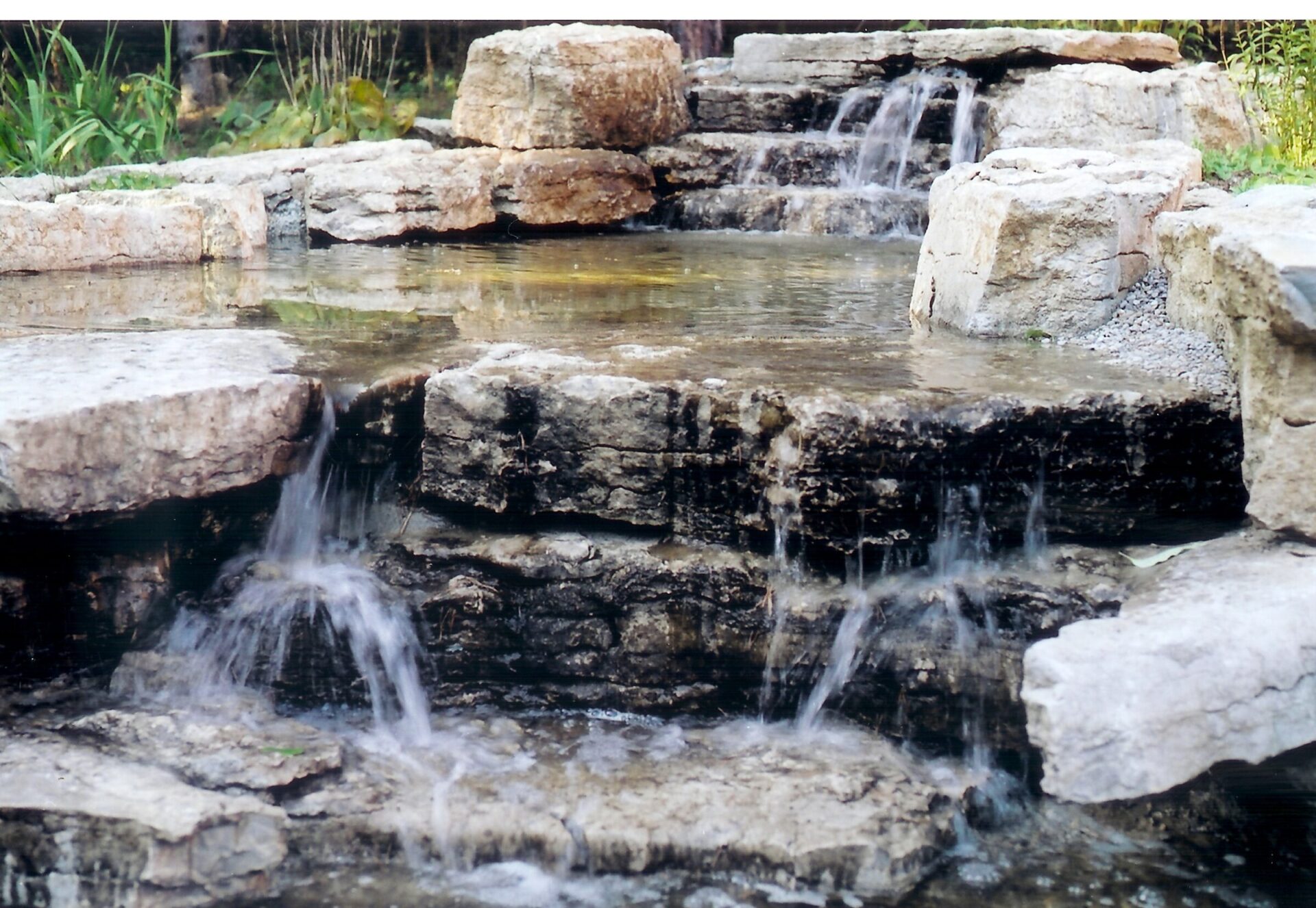 A small, multi-tiered waterfall flows over natural rocks, surrounded by greenery, creating a tranquil and peaceful natural scene.