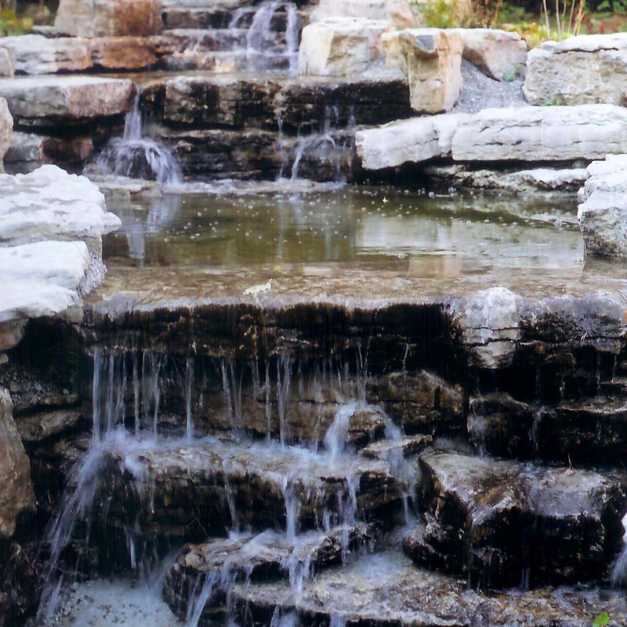 A tranquil, multi-tiered stone waterfall flows gently, surrounded by rocks and greenery, creating a serene natural landscape scene.