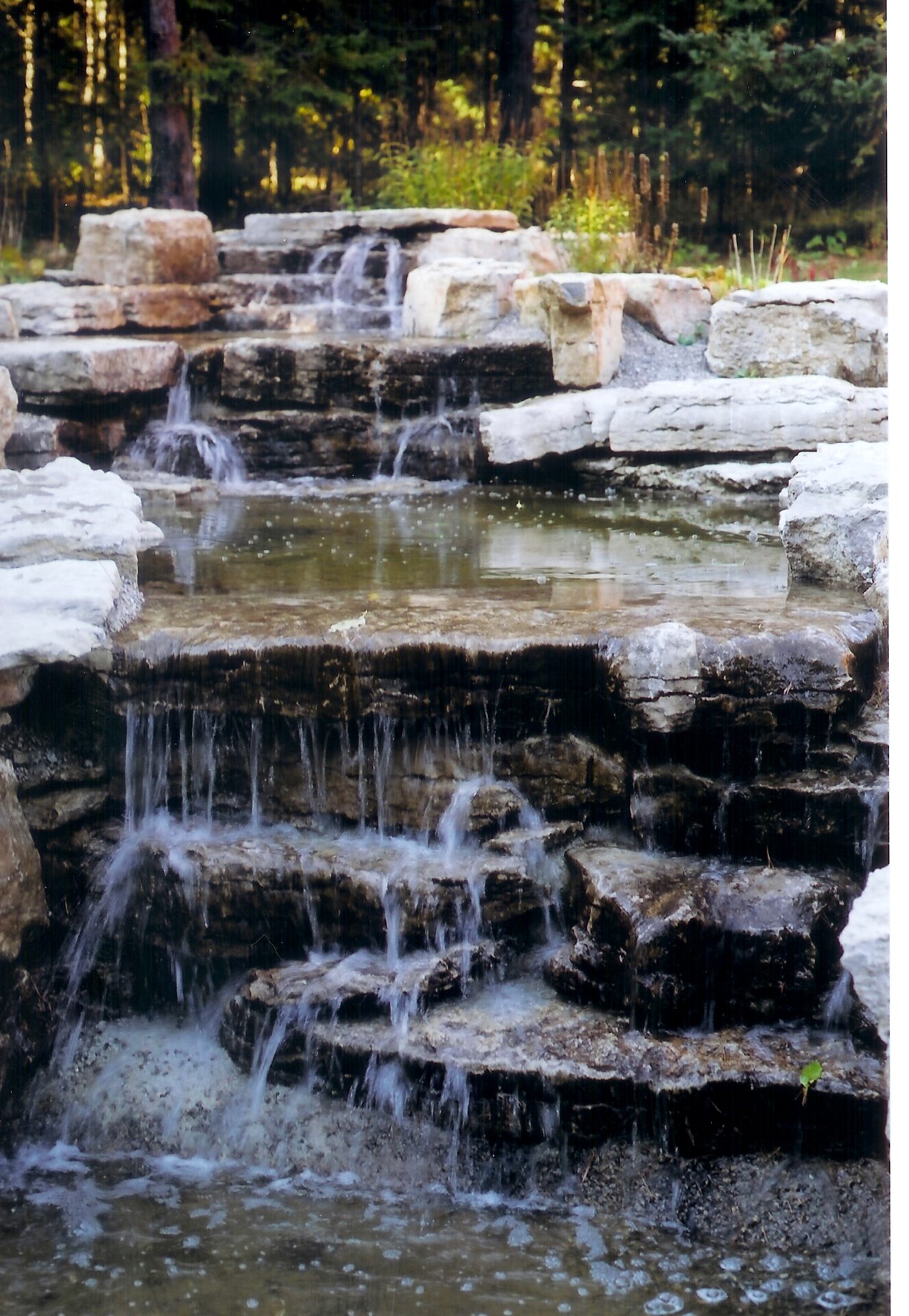 A small, multi-tiered waterfall cascades over rocks into a clear pool, surrounded by trees and foliage in a tranquil setting.