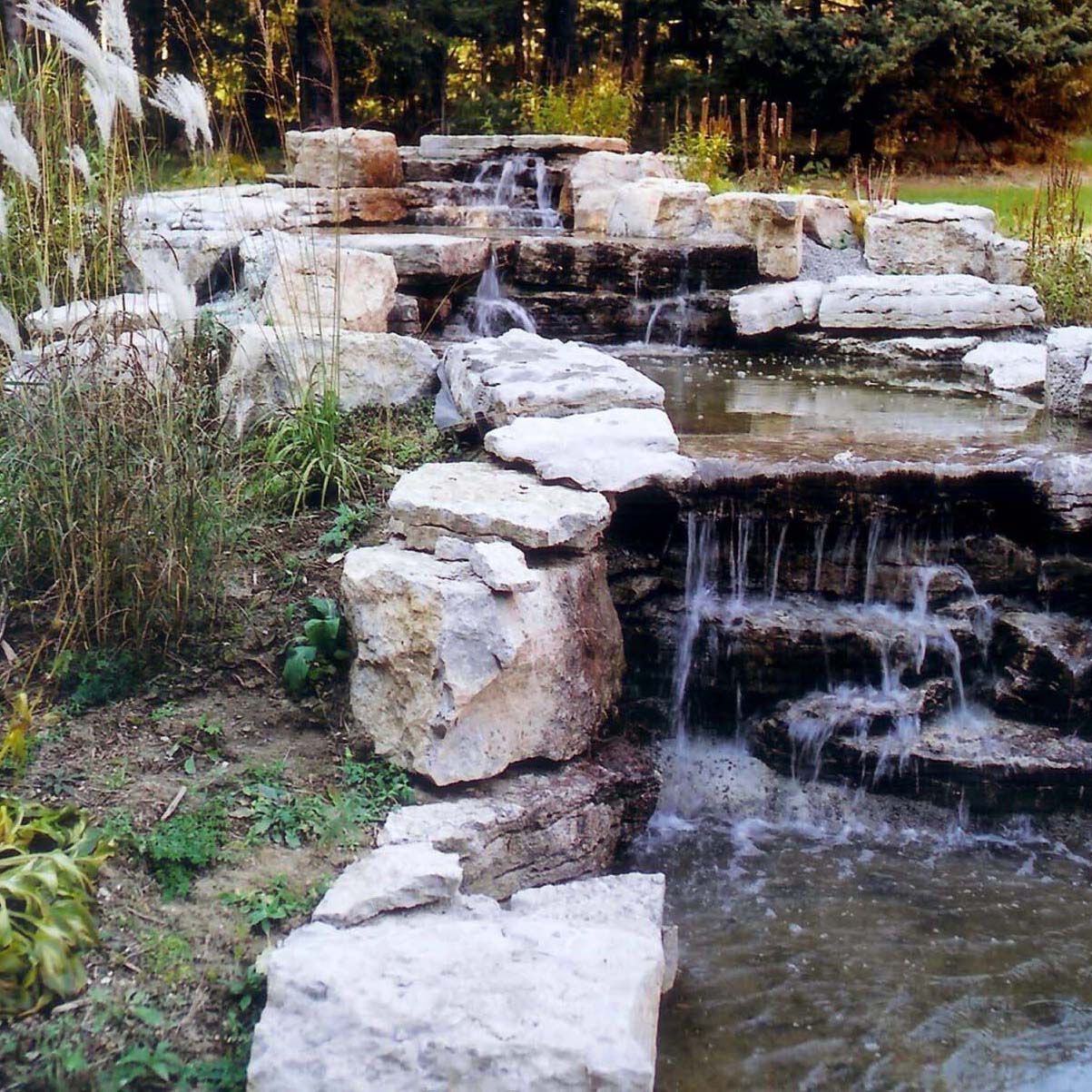 A serene garden scene with cascading water over layered stone formations, surrounded by lush greenery and natural vegetation. Tranquil outdoor setting.