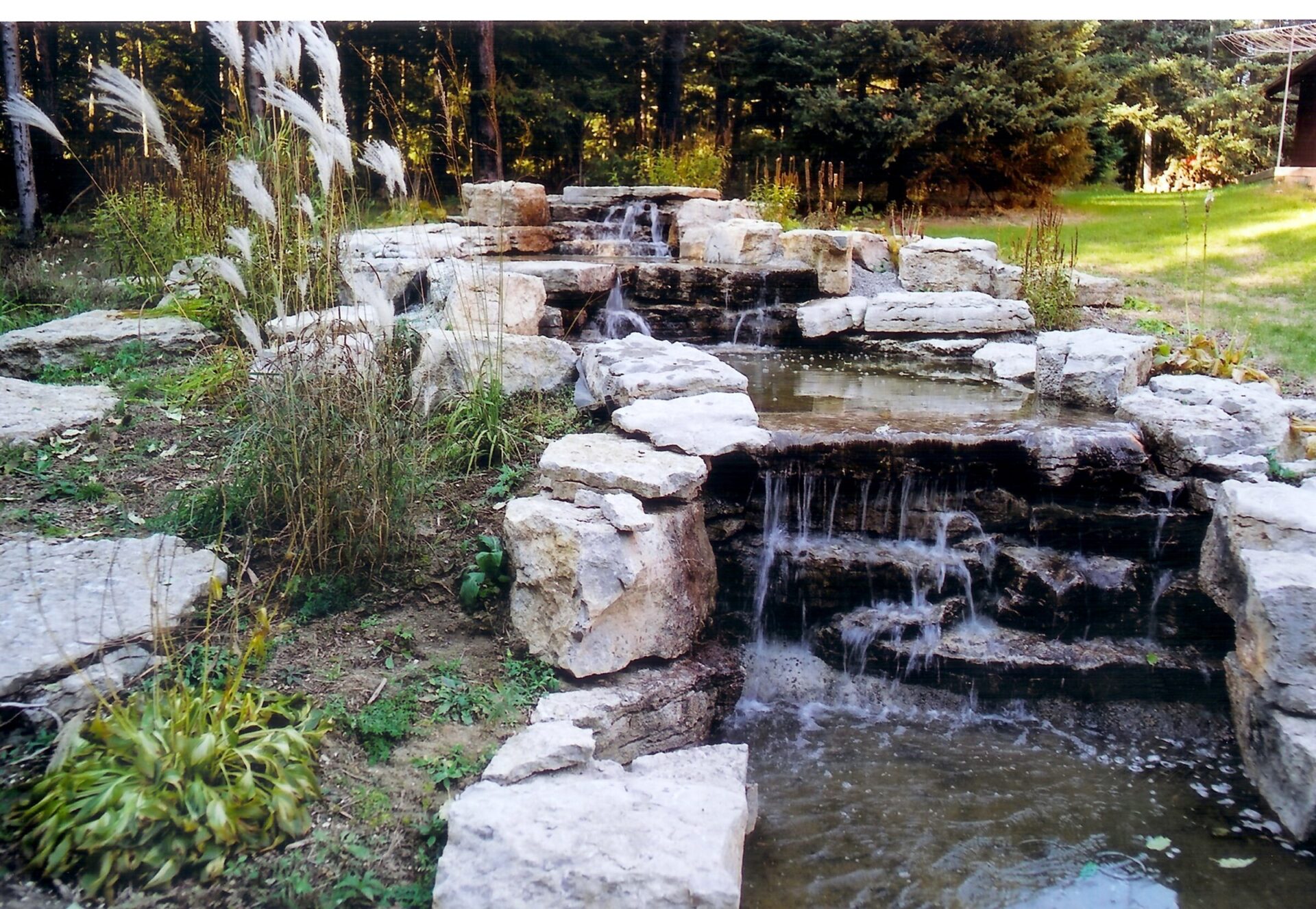 Rocky garden waterfall flows gently amidst greenery, creating a serene, natural scene. Surrounded by lush plants and bordered by tall trees.