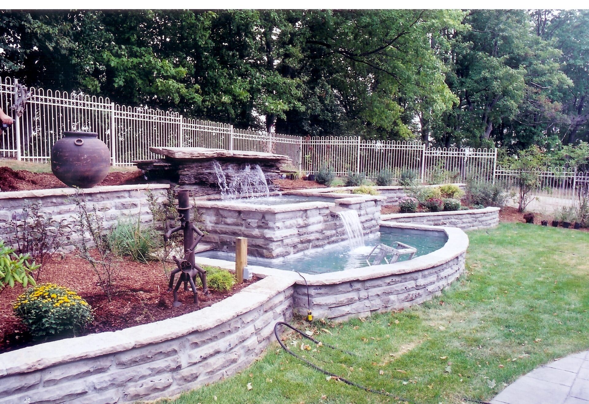 A landscaped garden features a tiered stone water fountain with cascading waterfalls, surrounded by greenery and a wrought iron fence.