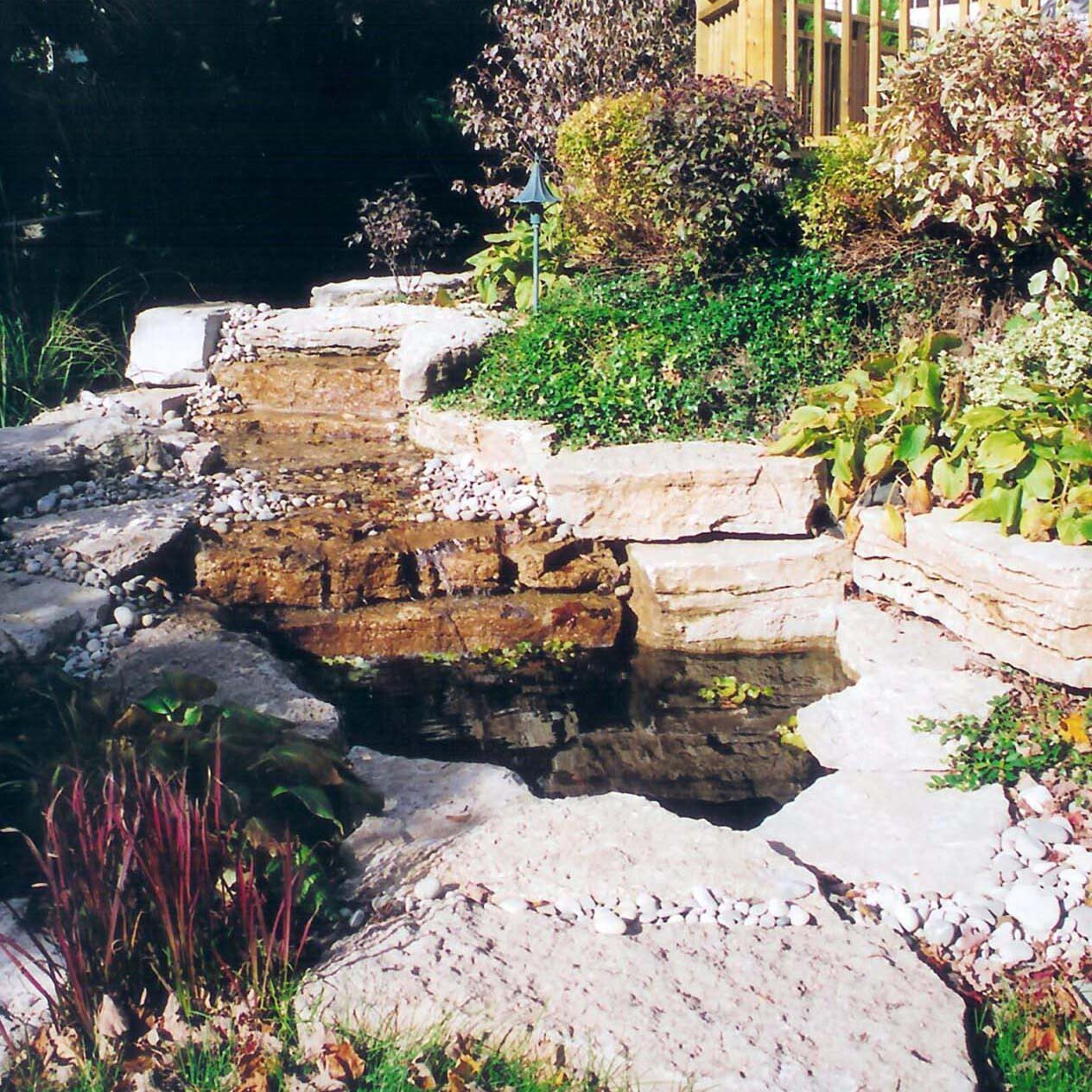 A small, tranquil garden features a cascading rock waterfall surrounded by lush greenery and colorful plants, creating a peaceful, natural setting.