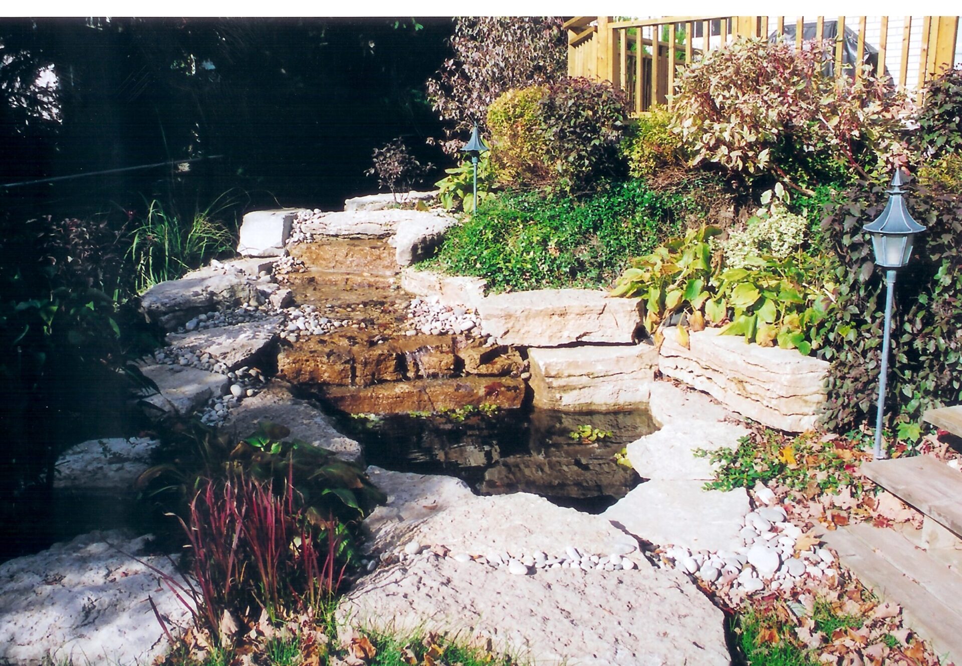 A serene garden with stone steps, a small waterfall, and lush greenery. A lamp post stands near a deck railing above.