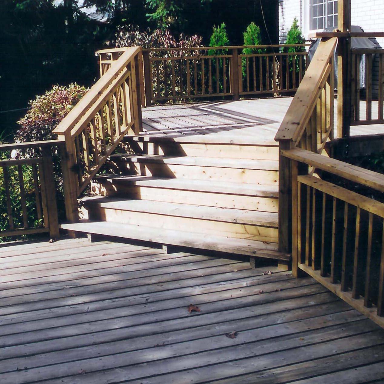 Wooden deck with stairs and railings, surrounded by greenery and a brick building. Sunlight casts shadows on the steps and platform.